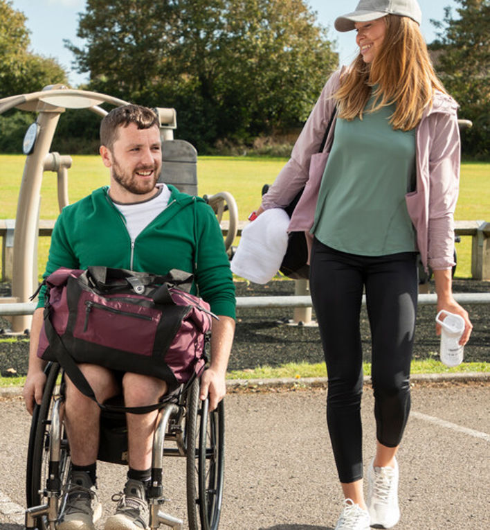 A woman walking with a man in a wheelchair, wearing sports gear, next to a car