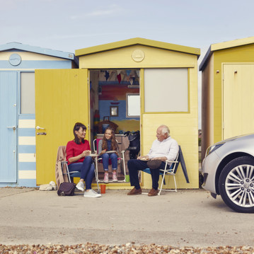An older man, a woman and a young girl sit outside a yellow beach hut, next to a car
