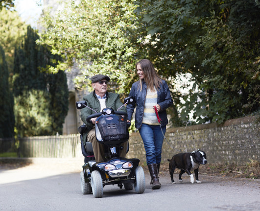 An older man riding a mobility scooter, next to a woman walking a dog