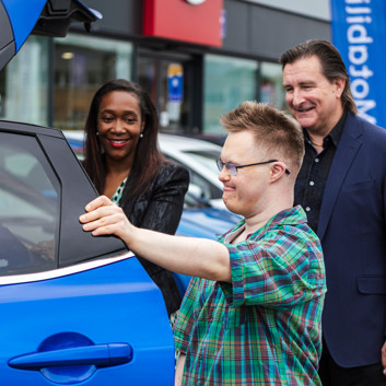 A young man looks inside a car at a dealer, with a smiling man and woman