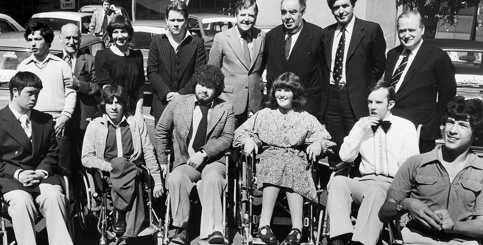 Six young people in wheelchairs pose for a black-and-white photo, with car dealers and Motability Operations staff