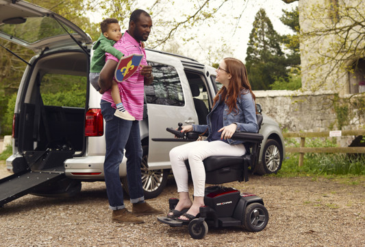 A man carrying a child on his back speaks to a woman in a wheelchair, next to a Wheelchair Accessible Vehicle