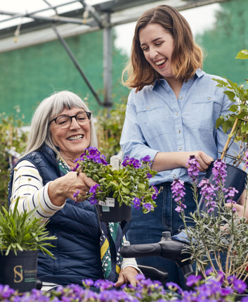 A woman on a mobility scooter shows another woman some flowers, in a garden centre