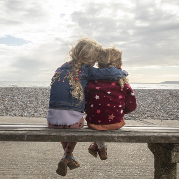 Two young girls sitting on a bench and hugging, looking at a beach