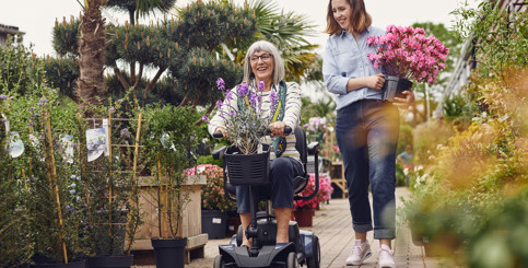 A smiling older woman on a mobility scooter, next to a smiling woman holding flowers
