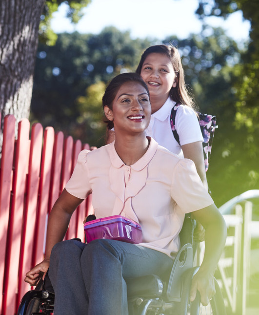 A smiling woman in a wheelchair with a lunch box, and a schoolgirl following behind her