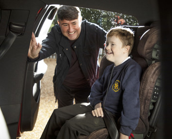 A smiling man helping a smiling school boy into his specially-adapted car seat