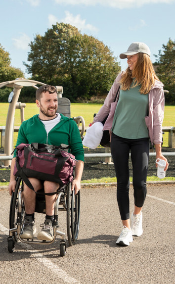 A woman walking with a man in a wheelchair, wearing sports gear, next to a car